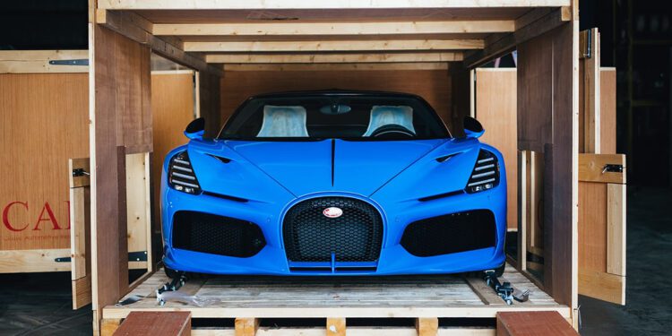 A Bugatti Mistral in striking French Racing Blue sits poised inside a wooden shipping crate, with its sleek front facing outward.