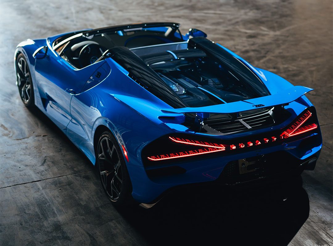 A sleek Bugatti convertible sports car, painted in iconic French Racing Blue, flaunts its modern design and LED taillights as it sits poised indoors on a concrete floor.