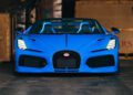 A Bugatti Mistral in stunning French Racing Blue with a convertible top is parked indoors, facing forward. The elegant sports car is framed by wooden crates, hinting at the showroom setting of Miller Motorcars.