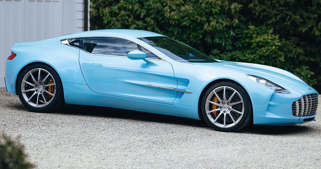 A sleek Aston Martin One-77 in Blue Moon Pearl is elegantly parked on a gravel driveway by a white garage, its presence commanding attention. In the background, trees whisper in the gentle breeze, enhancing the serene yet thrilling aura of this masterpiece from Miller Motorcars.