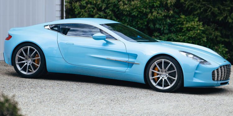 A sleek Aston Martin One-77 in Blue Moon Pearl is elegantly parked on a gravel driveway by a white garage, its presence commanding attention. In the background, trees whisper in the gentle breeze, enhancing the serene yet thrilling aura of this masterpiece from Miller Motorcars.