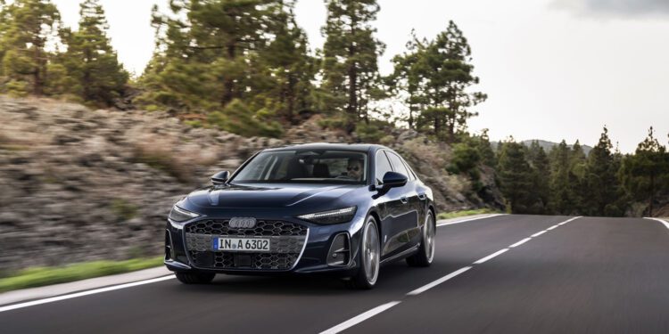 A dark blue new Audi A6 sedan glides along a winding, empty road bordered by trees and rocky terrain under a cloudy sky.