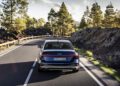 A dark blue Audi A6 sedan drives on a curving mountain road lined with trees and volcanic rocks under a partly cloudy sky.