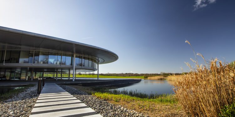 A sleek pathway leads to a modern, circular glass building by a serene pond under a clear blue sky with scattered clouds, symbolizing McLaren's brand reinvention—a fusion of innovation and elegance akin to their new future SUV.