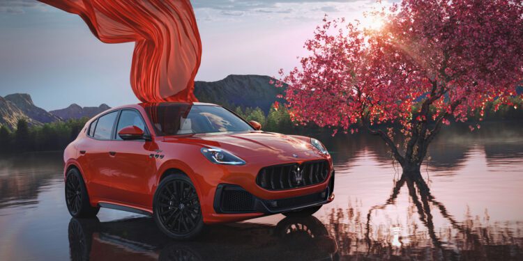 An orange 2025 Maserati Grecale SUV is parked beside a blooming pink tree near a reflective lake, with a red cloth draped over part of the car.