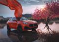 A red 2025 Maserati Grecale luxury SUV with a flowing red cloth above it is parked on a reflective water surface next to a blossoming tree at sunset.