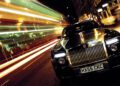A luxury Rolls-Royce with a prominent grille stands majestically on a city street at night, embodying the elegance of the Goodwood Era. Blurred light trails from passing vehicles craft a dynamic effect beside the car, highlighting its timeless allure.