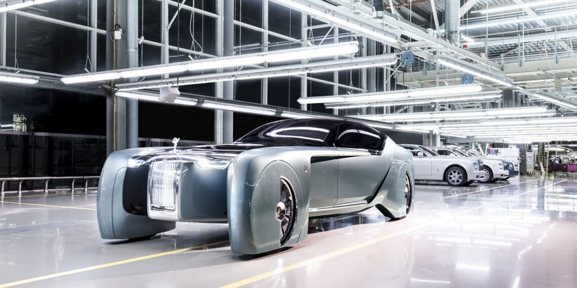 In an industrial showroom, the futuristic concept car stands out with its sleek design and large front grille, reminiscent of the Goodwood Era's elegance. Other cars linger in the backdrop, highlighting its innovation.