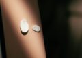 Two round cotton pads with lotion rest on a person’s skin, one larger than the other, against a blurred background reminiscent of cherry blossom petals in spring.