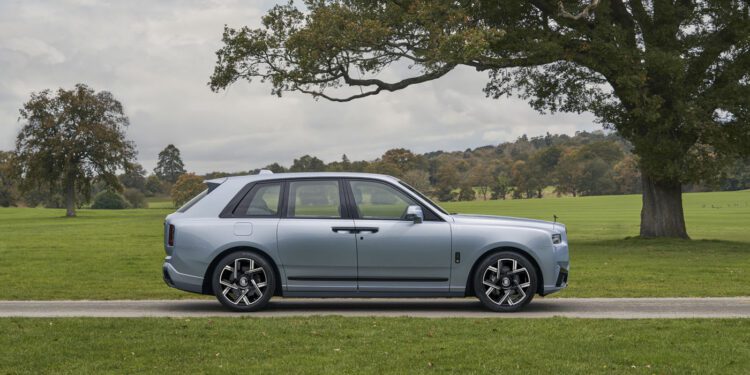 A light blue Rolls-Royce Cullinan is parked on a paved path in a grassy park with large trees and a cloudy sky in the background.