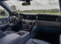 The Rolls-Royce Cullinan’s luxury interior features leather seats, a modern dashboard, touchscreen controls, and a refined steering wheel—all viewed from the front passenger side with a scenic landscape visible through the windows.