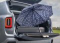 An open blue umbrella with a white daisy pattern is propped against the open trunk of a silver Rolls-Royce Cullinan in an outdoor setting.