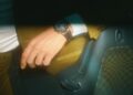 A person wearing a brown jacket and a Porsche Design Chronograph 911 wristwatch rests their hand on the seat of a car with patterned upholstery.