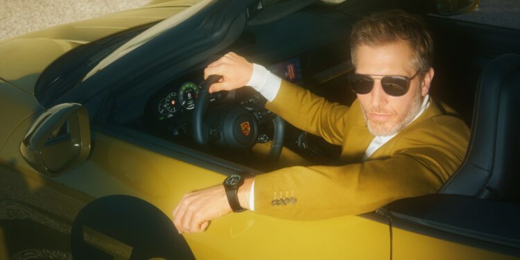 A man wearing sunglasses and a mustard-colored blazer sits in the driver’s seat of a gold convertible sports car, showcasing his Owner-Exclusive style and glancing toward the camera—his Porsche Design Chronograph 911 visible on his wrist.