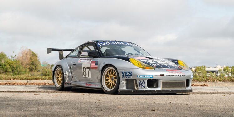 A sleek silver Porsche 911 GT3 RS, boasting the number 87 and adorned with Orbit Racing sponsor logos, is parked on a street lined with trees and buildings.