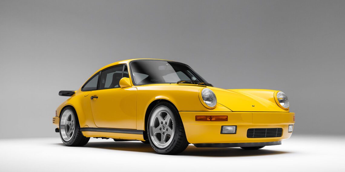 A 1989 RUF CTR Yellowbird, with its iconic yellow finish, two doors, circular headlights, and wide tires gleams under the bright studio lights. It's a striking piece of automotive history that would be a highlight at Broad Arrow Auctions.