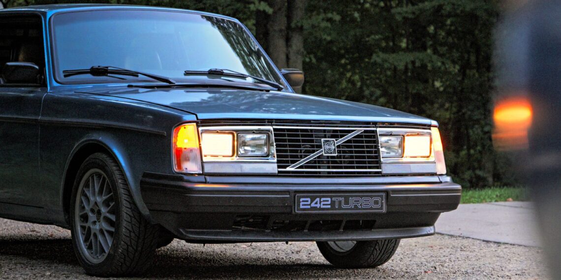 A classic Volvo 242 Turbo, reminiscent of its Group A Homologation days, with headlights on, parked on a gravel path against a backdrop of trees—a scene straight out of a Petrolicious story.