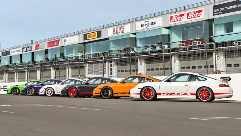Five Porsche sports cars, including a striking Porsche 911 GT3 RS, are parked in a row on a race track in front of a pit building with multiple sponsor signs.