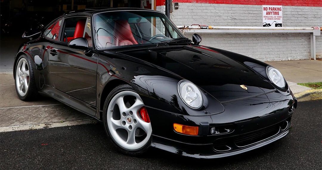 A black Porsche 911 Turbo with a striking red interior is parked in front of a building with a red and white brick wall and a "No Parking Anytime" sign, showcasing why it's one of the top Porsche models.