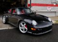 A black Porsche 911 Turbo with a striking red interior is parked in front of a building with a red and white brick wall and a "No Parking Anytime" sign, showcasing why it's one of the top Porsche models.