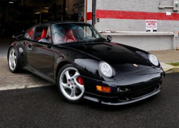 A black Porsche 911 Turbo with a striking red interior is parked in front of a building with a red and white brick wall and a "No Parking Anytime" sign, showcasing why it's one of the top Porsche models.