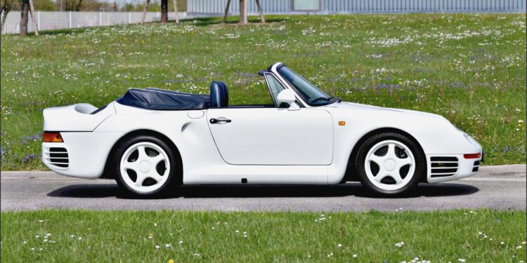 A white Porsche 959 Speedster with the top down is parked on a paved road next to a grassy field with scattered flowers, ready to turn heads at the RM Sotheby's Milan Auction.