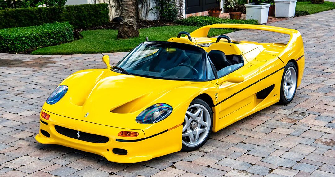 A Giallo Modena Ferrari F50 sports car is parked on a stone driveway in front of a landscaped garden and large house, exuding elegance worthy of a Ferrari auction.