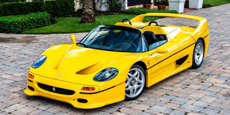 A Giallo Modena Ferrari F50 sports car is parked on a stone driveway in front of a landscaped garden and large house, exuding elegance worthy of a Ferrari auction.