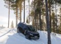 A black Range Rover Electric drives down a snowy slope surrounded by tall pine trees in Sweden, undergoing cold weather testing on a bright, sunny day.