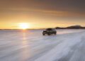 A black Range Rover Electric drives across a snowy, frozen landscape at sunset in Sweden, with mountains visible in the background during cold weather testing.