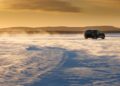 A dark Range Rover Electric drives across a snow-covered landscape in Sweden at sunset, braving blowing snow and distant hills—ideal conditions for cold weather testing.
