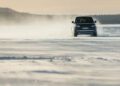 A Range Rover Electric drives through snow on a flat, open area in Sweden, cold weather testing with mist trailing behind under an overcast sky and distant tree line.