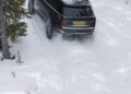 A Range Rover Electric with a yellow license plate drives uphill on a snowy, forested path in Sweden during cold weather testing.