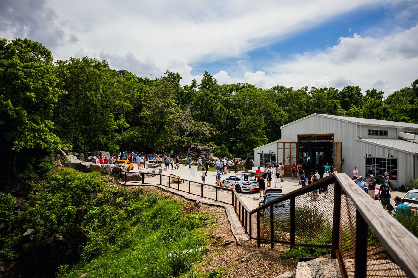 People gather outside a large metal building at a scenic location with trees and rocks under a partly cloudy sky, as sleek Porsches line up for the event.