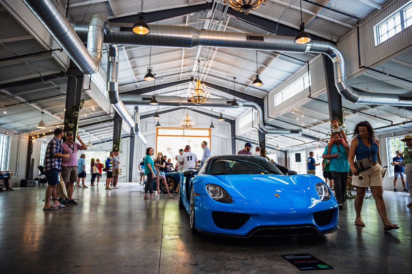 At a Porsche event in May, a stunning blue sports car was on display inside the large, industrial-style venue. Enthusiasts gathered around, capturing photos and engaging in passionate PLÁTICA as they admired the sleek vehicle.