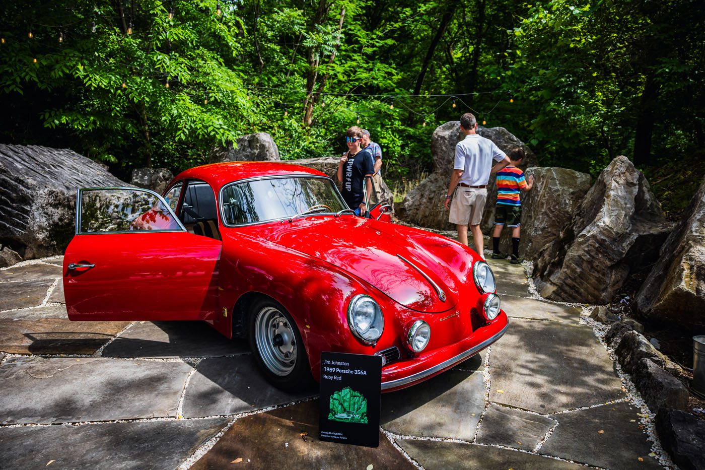 A red vintage Porsche with its driver's door open is showcased outdoors on stone paving, surrounded by people engaged in plática amidst lush greenery. An event sign detailing the car stands prominently in the foreground.