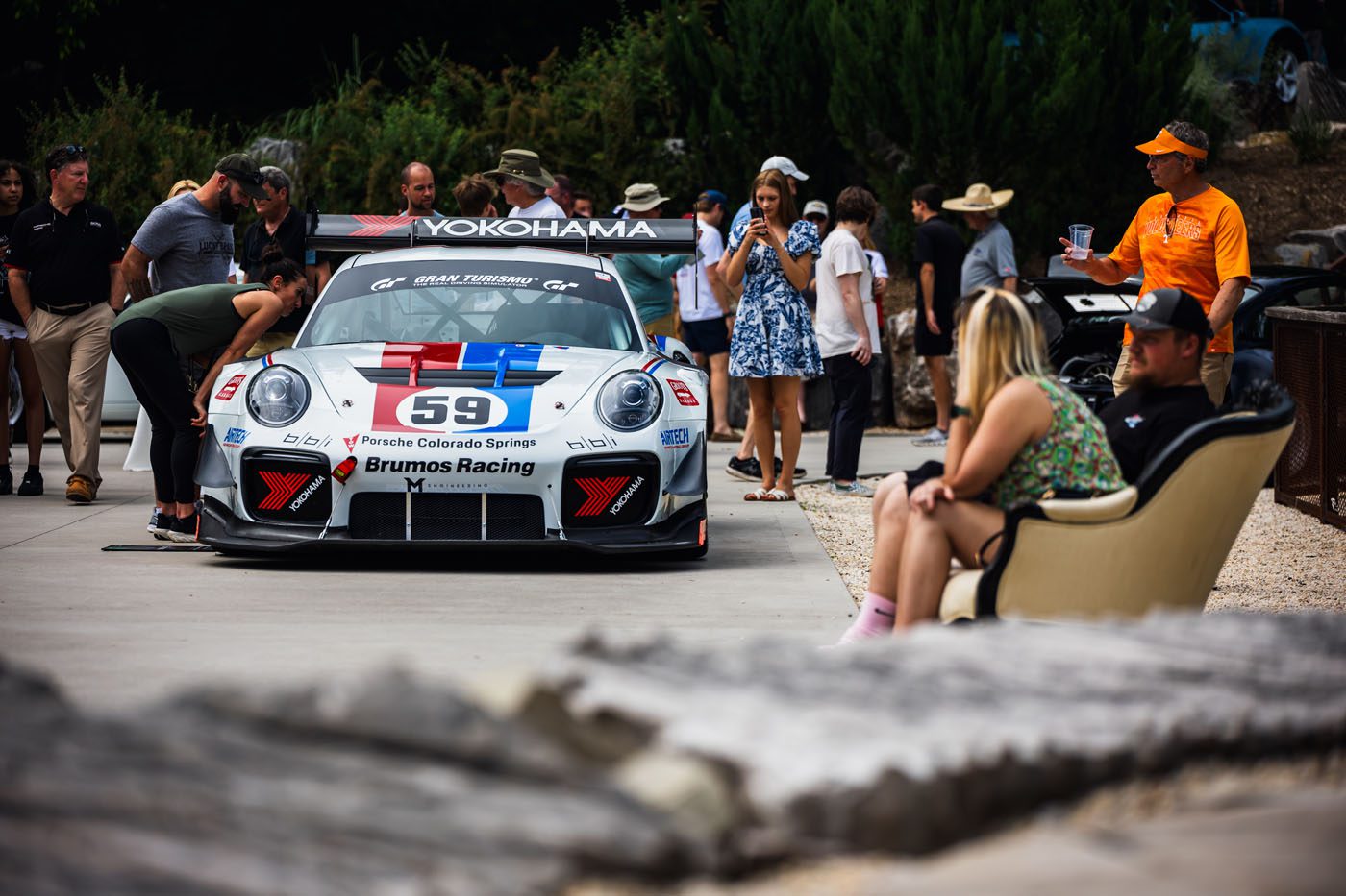 People gather around a parked Brumos Racing Porsche, number 59, under the sunny sky at the PLÁTICA event.