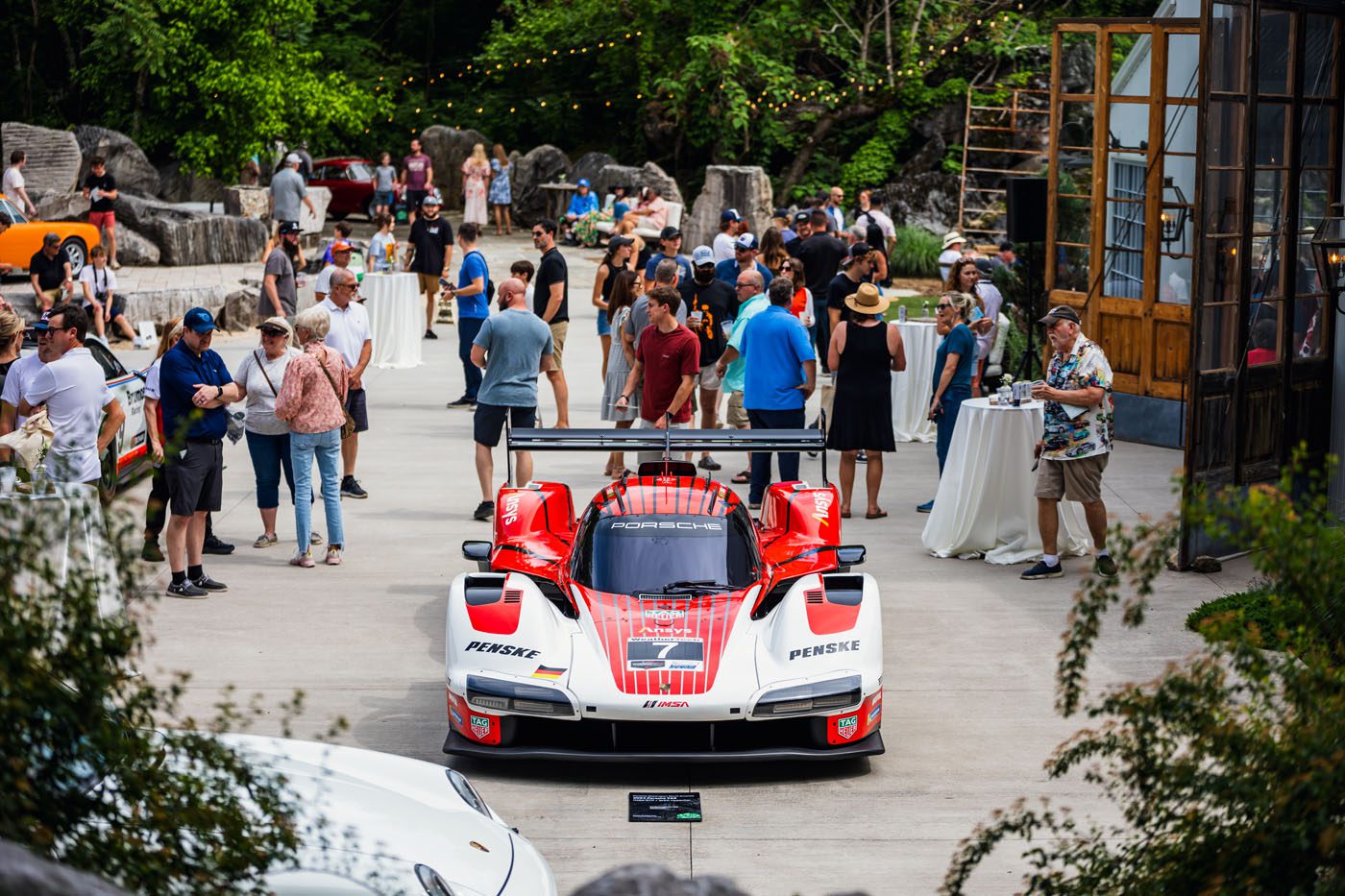 A red and white Porsche race car is displayed outdoors at an exciting event, surrounded by people and greenery, with tables and other cars in the background.