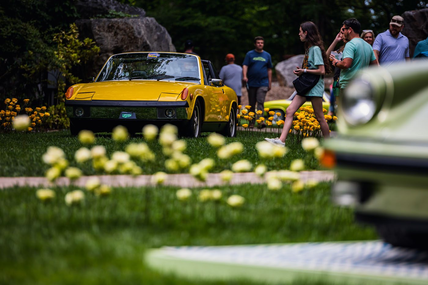 A yellow convertible car is displayed outdoors surrounded by flowers during the unforgettable Porsche event. People engage in lively plática as they walk around, while another car is partially visible in the foreground, adding to the vibrant atmosphere.