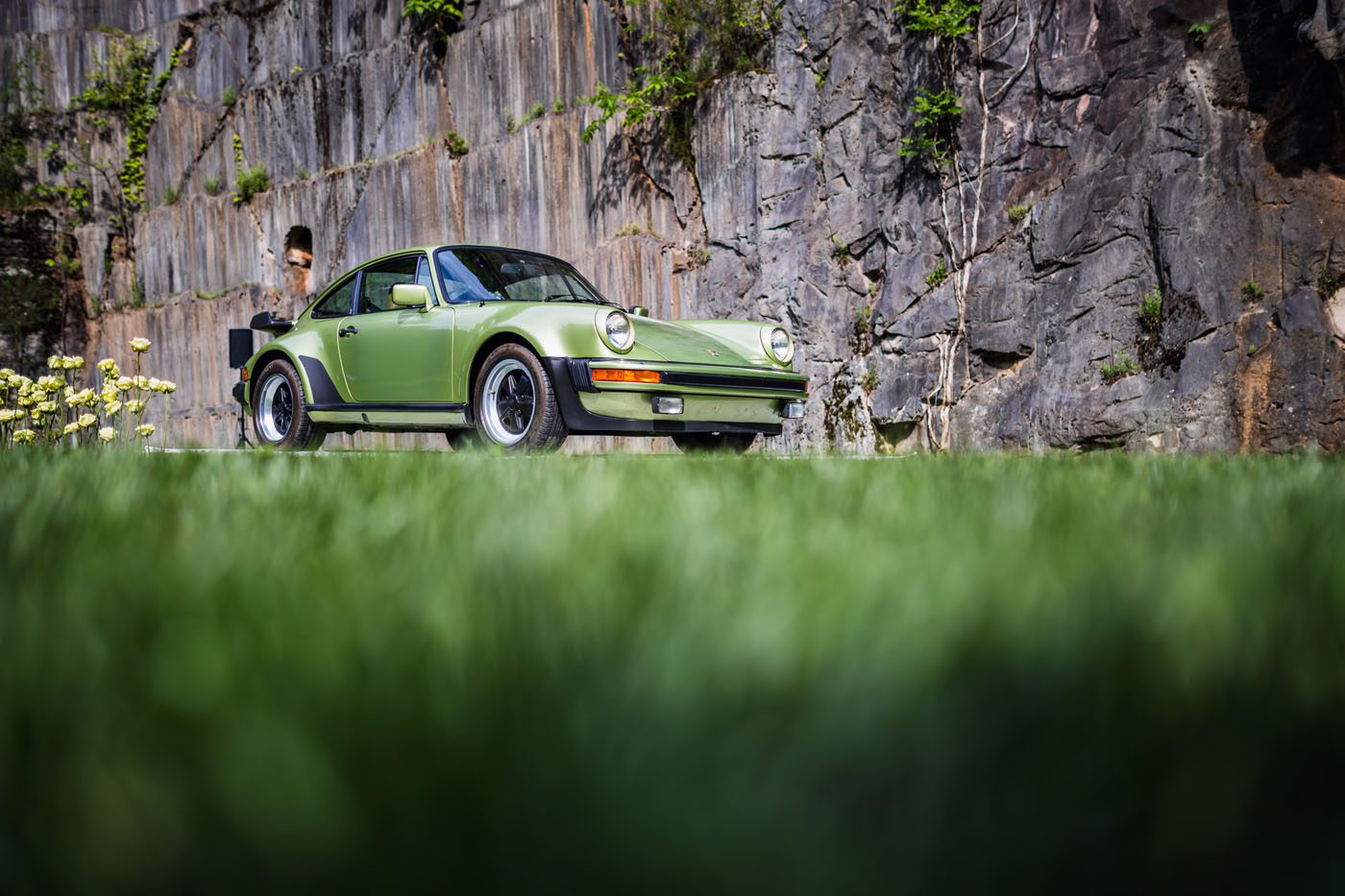 A green vintage car is elegantly parked on a grassy area with a rocky wall backdrop, capturing attention at the May Porsche event. Nearby, a person stands, perhaps engaging in PLÁTICA about the car's storied past.