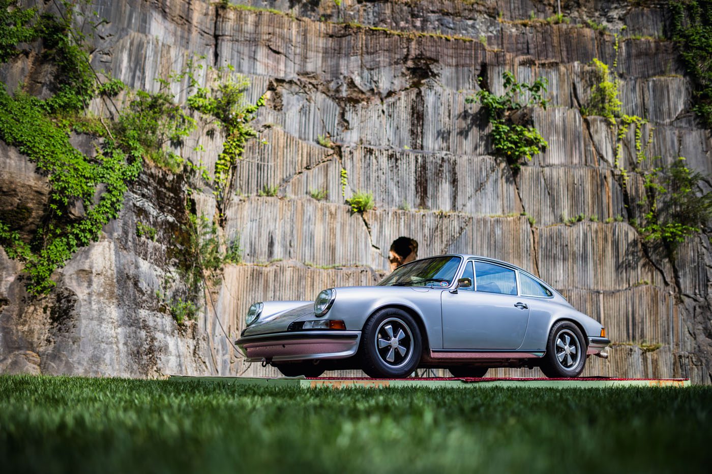 A classic silver Porsche is parked on the grass in front of a large stone wall, where greenery gently climbs, creating an ideal backdrop for any vintage car event.