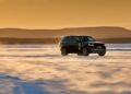 A Range Rover Electric SUV drives across a snow-covered landscape at sunset, with hills in the background and snow being kicked up behind the vehicle, showcasing its capability and impressive 300 mile range.