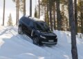 A Range Rover Electric SUV drives down a snowy, sloped trail surrounded by tall pine trees in a forested area, showcasing its 300 mile range.