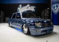 A custom blue van with silver flame graphics, low suspension, chrome wheels, and blue underglow lights is parked in an indoor garage at West Coast Customs, a legendary name in the world of custom cars.