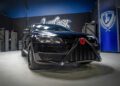 A black car customized with a Star Wars Darth Vader-themed front grill, inspired by custom cars at West Coast Customs, stands in a modern, well-lit garage with blue and black walls and tool cabinets.
