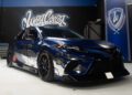 A dark blue custom race car with decals is parked indoors at West Coast Customs, surrounded by tool cabinets and a wall filled with logos—a true showcase of custom cars.