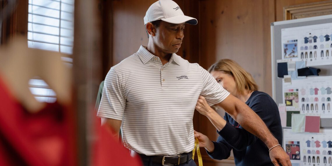 A man stands still while a woman measures his arm with a measuring tape in a wood-paneled room, as if preparing for a moment as memorable as getting your first car, with a design board visible in the background.