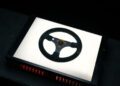 A black steering wheel, reminiscent of an F1 Victory moment, is displayed on a backlit white surface with red accents visible beneath.