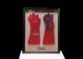 Framed display of a pair of red and gray gloves on a white background pays homage to Ayrton Senna’s F1 victory, set against a sleek black backdrop.