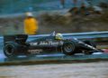 A black Lotus racing car with prominent "John Player Special" branding speeds down the track. The driver, perhaps reminiscent of Ayrton Senna, wears a helmet in the wet conditions; a trackside individual in yellow rain gear watches. Motion blur captures the essence of an F1 victory chase.